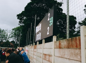 A large digital scoreboard at an outdoor sports venue displays a score of 2-2. Spectators are gathered, observing the event with interest. The scene is enclosed by a tall metal fence and surrounded by lush green trees, indicating a natural setting. Overcast skies add a muted tone to the environment.
