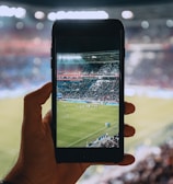 A hand holding a smartphone captures a live view of a soccer stadium filled with fans and players on the field. The image on the smartphone screen is crisp, showing a wide-angle view of the game in action.