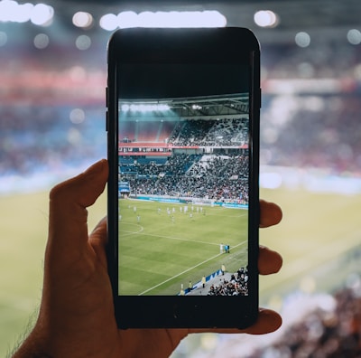 A hand holding a smartphone captures a live view of a soccer stadium filled with fans and players on the field. The image on the smartphone screen is crisp, showing a wide-angle view of the game in action.