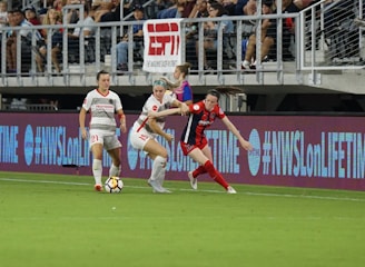 A competitive soccer match is underway with players vying for the ball near the sideline. The players wear different uniforms, indicating they are from opposing teams. The audience is visible in the stadium seating above, and there are visible advertisements and hashtags on the LED boards along the field.