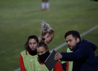 A group of individuals on a sports field are wearing green vests over red attire. One person is holding a tablet and seems to be discussing or showing something to the others. The atmosphere suggests a sports event, likely soccer, indicated by their clothing and the setting. The background is a grassy field.