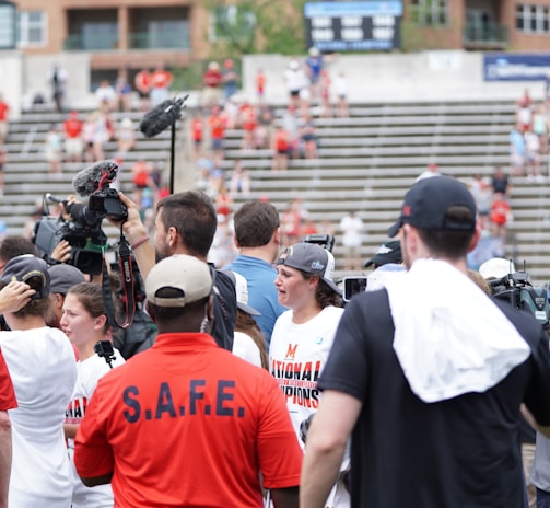 A lively gathering in an outdoor sports venue with a group of people, some wearing matching shirts labeled 'National Champions.' Several individuals are holding cameras and microphones, indicating a media presence. The background shows blurred spectators sitting on bleachers.