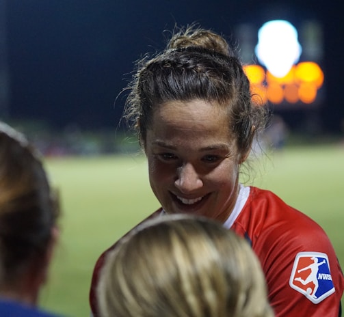 A female athlete in a red sports jersey with the NWSL logo energetically interacts with others on a sports field. The backdrop consists of a darkened night sky and a blurred scoreboard or advertisement display, with soft, colorful lights.