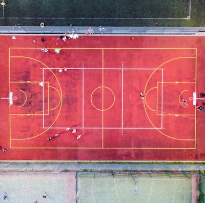 An aerial view of a sports court with a vibrant red surface and yellow lines marking different sections for basketball and soccer. Several people are present on the court, some engaged in activities, while others are standing along the perimeter. The court is enclosed by a metal fence and surrounded by grass on all sides.