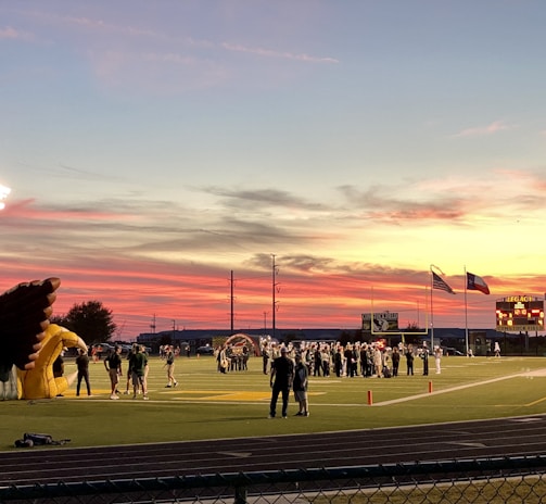 A sports field with a football game unfolding under a vibrant sunset sky, featuring a large inflatable mascot and a scoreboard displaying the score. Players and coaches are visible on the green field, surrounded by a track and some flags waving in the breeze.
