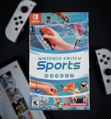 A box of Nintendo Switch Sports is prominently featured, surrounded by various Nintendo Switch controllers and a screen displaying a related interface. The box art depicts animated characters playing sports, such as tennis, with vibrant colors and action-oriented poses.