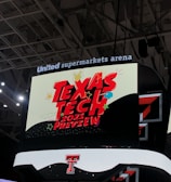 A scoreboard displaying the text 'Texas Tech 2021 Preview' in bold red letters with decorative elements like stars and clouds. The background is predominantly dark with bright accent colors, and the setting is within an indoor sports arena, specifically the United Supermarkets Arena.