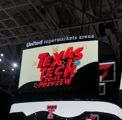 A scoreboard displaying the text 'Texas Tech 2021 Preview' in bold red letters with decorative elements like stars and clouds. The background is predominantly dark with bright accent colors, and the setting is within an indoor sports arena, specifically the United Supermarkets Arena.