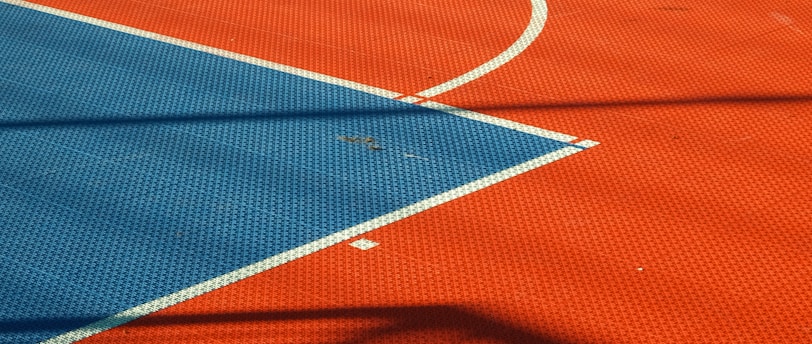 A vibrant sports court with sections of red and blue, featuring crisp white lines marking the boundaries. Shadows from a structure create geometric patterns across the surface, adding depth and interest to the scene.