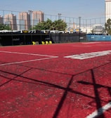 A sports court with a red playing surface and white lines, surrounded by a black mesh fence. In the background, tall buildings are visible, along with some trees and a few yellow chairs positioned near the fence.