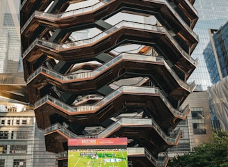 A large, spiral architectural structure with multiple levels stands prominently in an urban setting. In front of the structure, a large screen displays a sports broadcast, and several people are gathered around, some sitting and others standing. The scene is set outdoors, with towering modern buildings surrounding the area. The ground is paved with a geometric pattern of gray and white tiles.